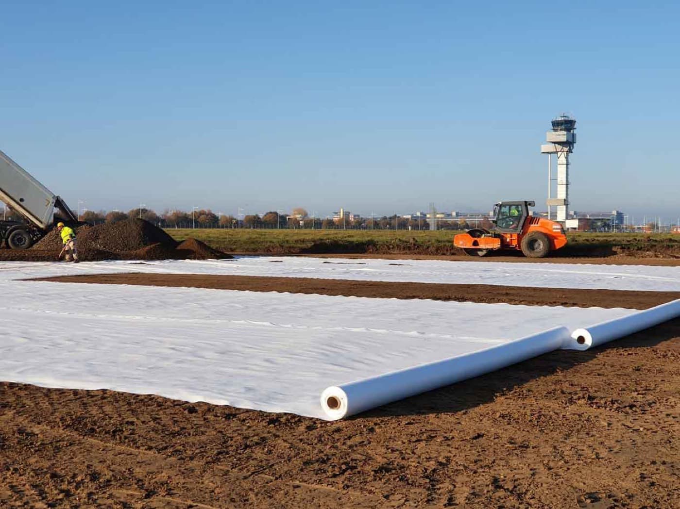 A wide roll of white non-woven geotextile being unrolled over a prepared subgrade, with a compactor working alongside.