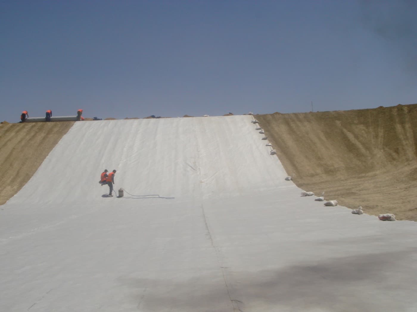 Geotextile laid across a long embankment slope, ballasted at the edges, with two workers on the fabric.